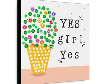 May include: A square wall art print featuring a yellow polka-dot pot containing a green topiary with colorful small flowers. To the right, the text "YES girl Yes" is printed in a black typewriter-style font against a white background.
