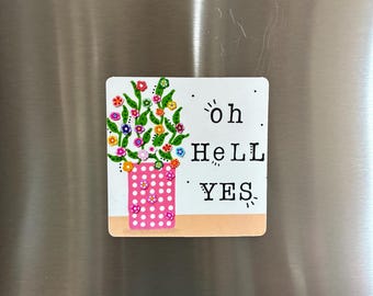 May include: A square magnet featuring a pink polka-dot vase filled with green stems and colorful small flowers. To the right of the vase, the text reads "oh HeLL YES" in black, stylized lettering. The background is white with a tan base.