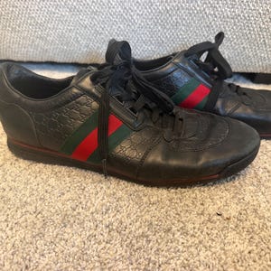 Puede incluir: Un par de zapatillas de cuero negro con un patrón de monograma en relieve, cordones negros y un detalle de banda lateral a rayas rojas y verdes. El calzado de corte bajo incluye suelas negras y punteras redondeadas.