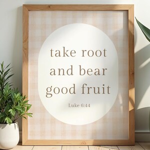 Könnte beinhalten: Holzgerahmtes Wandbild mit dem Text "take root and bear good fruit" in einem weißen Oval. Der Hintergrund hat ein hellbraunes Vichy-Muster. Das Kunstwerk wird von grünen Topfpflanzen flankiert.