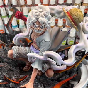 Peut inclure: Figurine détaillée d'un personnage aux cheveux blancs et hérissés, avec un chapeau de paille et un large sourire. La figurine est dans une pose dynamique, entourée d'effets d'énergie orange et blanche flamboyants, sur une base rocheuse. Le personnage porte une tenue gris clair.