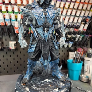 Puede incluir: Figura detallada de un personaje con armadura azul y negra con capucha. La figura está sobre una base texturizada con detalles helados. La figura tiene detalles intrincados y está rodeada de materiales de pintura.