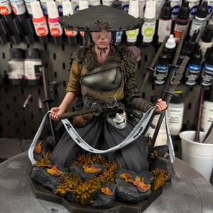 以下が含まれることがあります： 伝統的な日本の衣装を着た女性戦士の精巧なフィギュア。広い麦わら帽子と刀が含まれています。白い布とマスクでポーズをとっています。ベースには岩と紅葉があります。