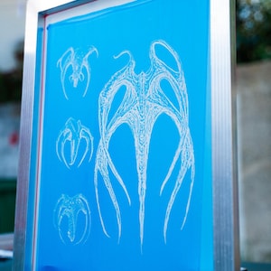 Peut inclure: Un cadre de sérigraphie avec une surface bleu vif. L'écran présente une illustration blanche stylisée d'une créature avec de multiples appendices. Le cadre est en métal argenté et est incliné vers le spectateur.