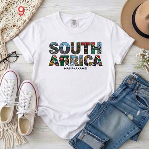 May include: A white short-sleeve t-shirt featuring the text SOUTH AFRICA in large block letters, with each letter filled with colorful illustrations of landmarks, wildlife, and cultural symbols, and the word MASIPHAKAME! printed below.
