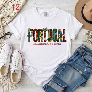 May include: A white short-sleeve t-shirt featuring the word PORTUGAL in large, colorful, patterned block letters. Each letter contains illustrations of Portuguese landmarks and symbols. Below the text is the phrase CORA&Ccedil;&Atilde;O DE LE&Atilde;O, ALMA DE CAMPE&Atilde;O.