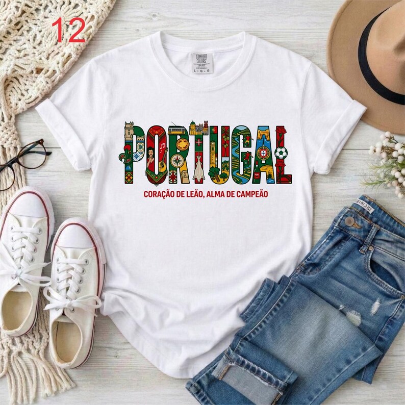 May include: A white short-sleeve t-shirt featuring the word PORTUGAL in large, colorful, patterned block letters. Each letter contains illustrations of Portuguese landmarks and symbols. Below the text is the phrase CORA&Ccedil;&Atilde;O DE LE&Atilde;O, ALMA DE CAMPE&Atilde;O.