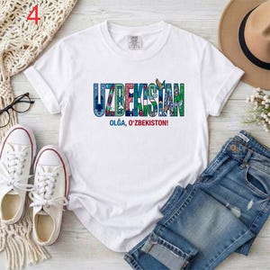 May include: A white short-sleeved t-shirt featuring the word UZBEKISTAN in large, multi-colored letters filled with intricate patterns and cultural motifs. Below the main text, the phrase OLĞA, O'ZBEKISTON! is printed in blue and red lettering.