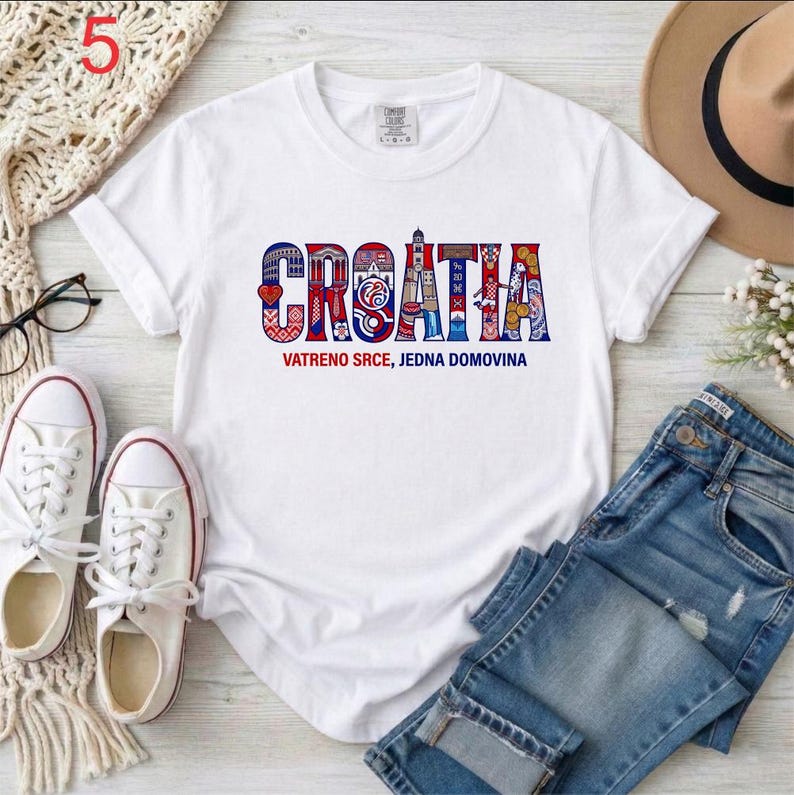 May include: A white short-sleeved t-shirt featuring the word CROATIA in large, stylized letters filled with red, white, and blue patterns. Below the word, the text reads VATRENO SRCE, JEDNA DOMOVINA in smaller blue and red font on the chest.