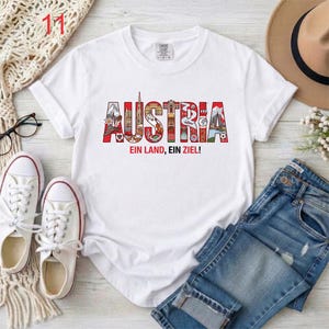 May include: A white short-sleeved t-shirt featuring the word AUSTRIA in large, colorful letters filled with various illustrations of landmarks and cultural symbols, with the text EIN LAND, EIN ZIEL! printed in red and black below the word.