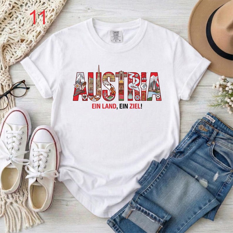 May include: A white short-sleeved t-shirt featuring the word AUSTRIA in large, colorful letters filled with various illustrations of landmarks and cultural symbols, with the text EIN LAND, EIN ZIEL! printed in red and black below the word.