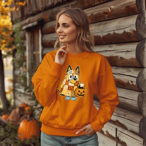 May include: An orange crewneck sweatshirt featuring a graphic of a blue dog character wearing a patchwork ghost costume and holding a jack-o'-lantern pumpkin. The sweatshirt has long sleeves, a ribbed neckline, and ribbed cuffs at the wrists.