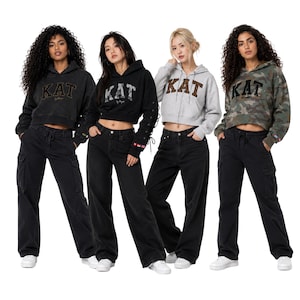 May include: Four cropped hoodies featuring the text KAT in various patterns, paired with black cargo or wide-leg jeans. The hoodies include a charcoal grey, a black with lace-up sleeves, a light grey zip-up, and a camouflage print design.