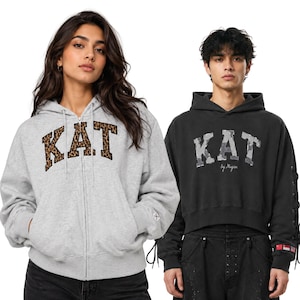 Può includere: Una felpa con zip grigio chiaro con applicazione KAT leopardata e una felpa nera con cappuccio con applicazione KAT mimetica, entrambe con maniche lunghe, cappucci con coulisse e polsini a coste su uno sfondo bianco uniforme.
