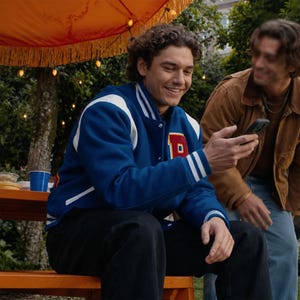 May include: A blue and white letterman jacket with a red "P" patch. The jacket is worn by a person sitting on a wooden bench. The person is smiling and looking at a smartphone. Another person is standing nearby.