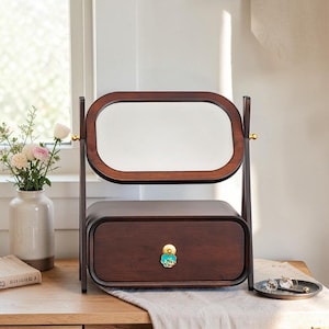 May include: A dark brown wooden vanity mirror with a rectangular frame and rounded corners. The mirror is mounted on a stand above a single storage drawer. The drawer features a decorative, circular, turquoise and gold-coloured metal pull handle.