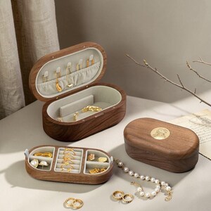 May include: Wooden jewelry boxes in various sizes and shapes, some open to display gold jewelry. The boxes are oval and rectangular, with a light-colored interior. A closed box features a gold emblem on the lid. A pearl necklace and gold rings are also visible.