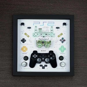 May include: A framed PlayStation 2 controller and components display. The black frame surrounds a white background with a disassembled controller, buttons, and circuit board. The PlayStation 2 and Sony logos are visible.