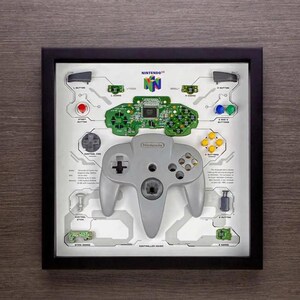 以下が含まれることがあります： Nintendo 64 コントローラーとコンポーネントの額入りディスプレイ。中央に灰色のコントローラーがあり、ラベル付きのパーツと緑色の回路基板に囲まれています。任天堂のロゴが見えます。フレームは黒です。