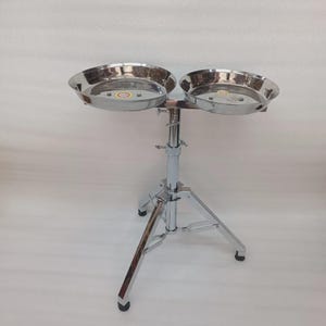 May include: A chrome-plated percussion instrument stand featuring two circular, stainless steel bowls. The bowls are mounted on an adjustable stand with a tripod base, designed for stability. The stand is suitable for holding percussion instruments.