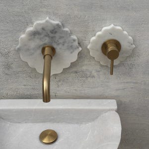Puede incluir: Un lavabo de baño con un lavabo de mármol blanco y accesorios a juego. El grifo y el control del agua son dorados, montados en placas traseras decorativas de mármol. El lavabo tiene una tapa de desagüe dorada.