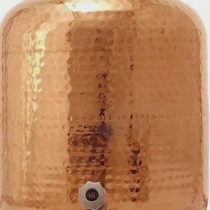 May include: A copper water container with a hammered texture. The container has a rounded body, a lid with a small handle, and a spigot near the base. The copper has a warm, reflective finish. The container is likely used for storing or dispensing water.