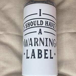 Puede incluir: Vaso blanco con tapa negra. El vaso tiene el texto negro "I SHOULD HAVE A (WARNING) LABEL". El vaso es cilíndrico y parece ser de metal.