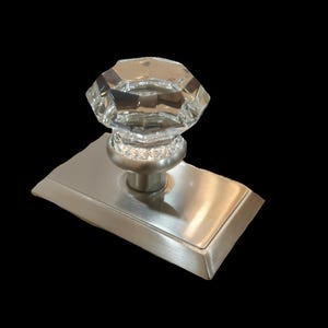 May include: A clear, multi-faceted, octagonal glass knob mounted on a brushed nickel rectangular backplate. The knob features a decorative ring of small, clear crystals at its base, connecting the glass top to the metallic hardware finish.