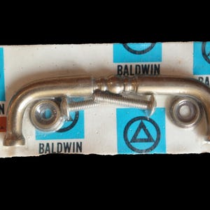 May include: A vintage Baldwin plumbing part, packaged on a white and blue card. The metal part is curved with a screw and nut, and the card has the Baldwin logo and text.