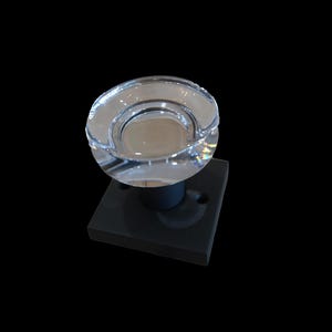 May include: A clear, faceted glass knob with a circular design, mounted on a black square base. The knob has a reflective surface, and the base has two screw holes. The background is solid black.