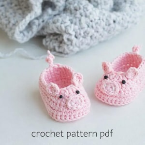 Peut inclure: Deux chaussons de bébé roses en crochet faits à la main, en forme de cochons, avec des yeux noirs et de petites oreilles. Ils sont posés sur une surface blanche, avec une couverture en crochet grise en arrière-plan. Le texte "crochet pattern pdf" est en bas.