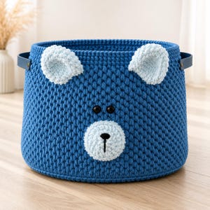 Peut inclure: Un panier de rangement rond en crochet bleu foncé représentant un visage d'ours. Le panier possède deux oreilles en crochet bleu clair, deux petits yeux en boutons noirs et un museau en crochet bleu clair avec un nez brodé en noir. Il a deux anses latérales bleu foncé.