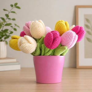May include: A bouquet of crocheted tulips in shades of yellow, cream, light pink, and magenta, featuring green crocheted stems and leaves. The flowers are arranged in a solid, smooth, bright pink cylindrical pot with a flat base and rim.