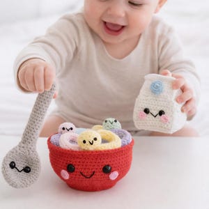 May include: A crocheted toy set featuring a red bowl filled with colorful cereal, a gray spoon, and a white milk carton. The bowl, spoon, and milk carton have smiling faces. The set is designed to look like a bowl of cereal.
