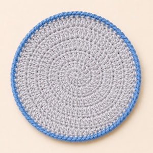 Peut inclure: Un dessous de verre circulaire au crochet présentant un centre gris clair travaillé en spirale, terminé par une bordure bleu vif. L'article textile est construit avec des mailles serrées et uniformes, créant une texture de surface plate.