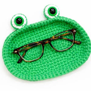 Peut inclure: Un plateau en crochet vert vif fait à la main en forme de grenouille, avec deux yeux blancs et verts. Des lunettes à monture écaille reposent à l'intérieur du plateau, sur fond blanc. Un article fantaisiste et fonctionnel.