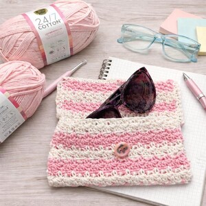 Peut inclure: Une pochette à lunettes crochetée à rayures roses et blanches, avec une fermeture à bouton. Des lunettes de soleil sont à l'intérieur. On aperçoit du fil rose et un crochet. Un carnet, un stylo et des lunettes sont également présents.