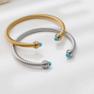Two twisted cable cuff bracelets, one gold and one silver, featuring turquoise beads at each end, resting on a white surface.