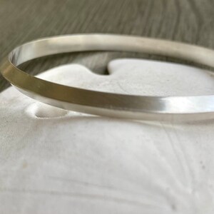 May include: A silver bangle bracelet with a flat, beveled edge. The bracelet is smooth and shiny.