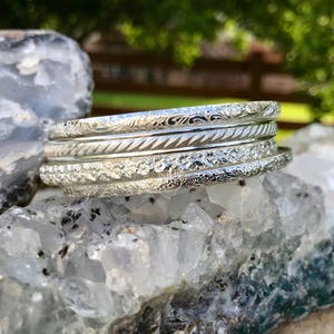 May include: A set of four silver bangles with intricate floral and geometric designs. The bangles are stacked on top of each other and are resting on a bed of white crystals.