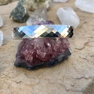 May include: A silver bracelet with a faceted, geometric design. The bracelet is resting on a cluster of purple crystals.