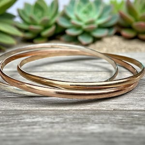 May include: Three thin bangles in silver, rose gold, and gold tones are stacked on a weathered wooden surface. The bracelets are smooth and circular, reflecting light. Succulents are visible in the background.