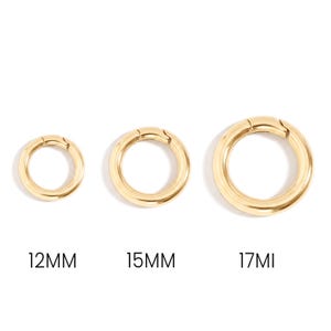 Könnte beinhalten: Drei goldfarbene Metall-Federringe in einer Reihe, beschriftet mit den Größen 12mm, 15mm und 17mm. Jeder kreisförmige Verschluss verfügt über einen Klappmechanismus, eine glatte, polierte Oberfläche und einen einheitlichen Goldton.