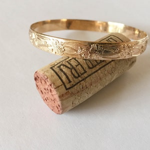 May include: A gold bangle bracelet with a floral design. The bracelet is on top of a wine cork.