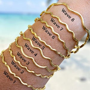 May include: Eight gold-toned metal bangles with a wavy, undulating design. Each bangle is labeled with black text ranging from Wave 1 to Wave 8, indicating the specific style of the wavy pattern on each individual piece of jewelry.