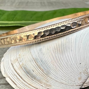 May include: A gold-colored bangle bracelet with a hammered texture and a detailed border. The bracelet is displayed on a white seashell, showcasing its design and craftsmanship. Jewelry.