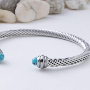 A close-up view of a silver twisted cable cuff bracelet featuring a turquoise stone end against a blurred background of stones.