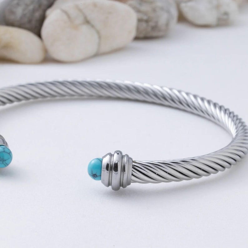 A close-up view of a silver twisted cable cuff bracelet featuring a turquoise stone end against a blurred background of stones.