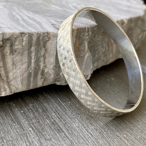 May include: A silver-colored bangle bracelet with a woven pattern. The bracelet has a smooth, polished interior and a textured exterior. The design is a repeating pattern of interwoven lines, creating a basket-weave effect. The jewelry is displayed on a gray stone surface.