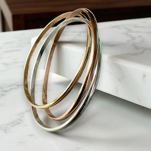 May include: A set of four metal bangles in gold, silver, and rose gold tones. The bangles are smooth and round, with a polished finish. The jewelry is displayed on a white marble surface.